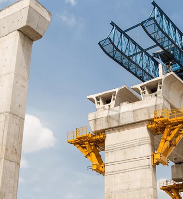 Large concrete pillars and elevated bridge segments under construction with yellow scaffolding and a blue overhead gantry crane against a partly cloudy sky.